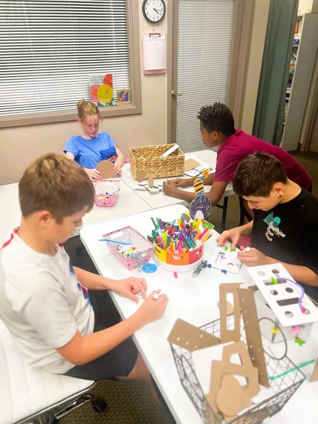 Four upper-elementary students sit at a table covered with cardboard pieces, connectors, and art materials. Each student is cutting or assembling small components, working independently with focused attention during the early preparation phase of the habitat-building task.
