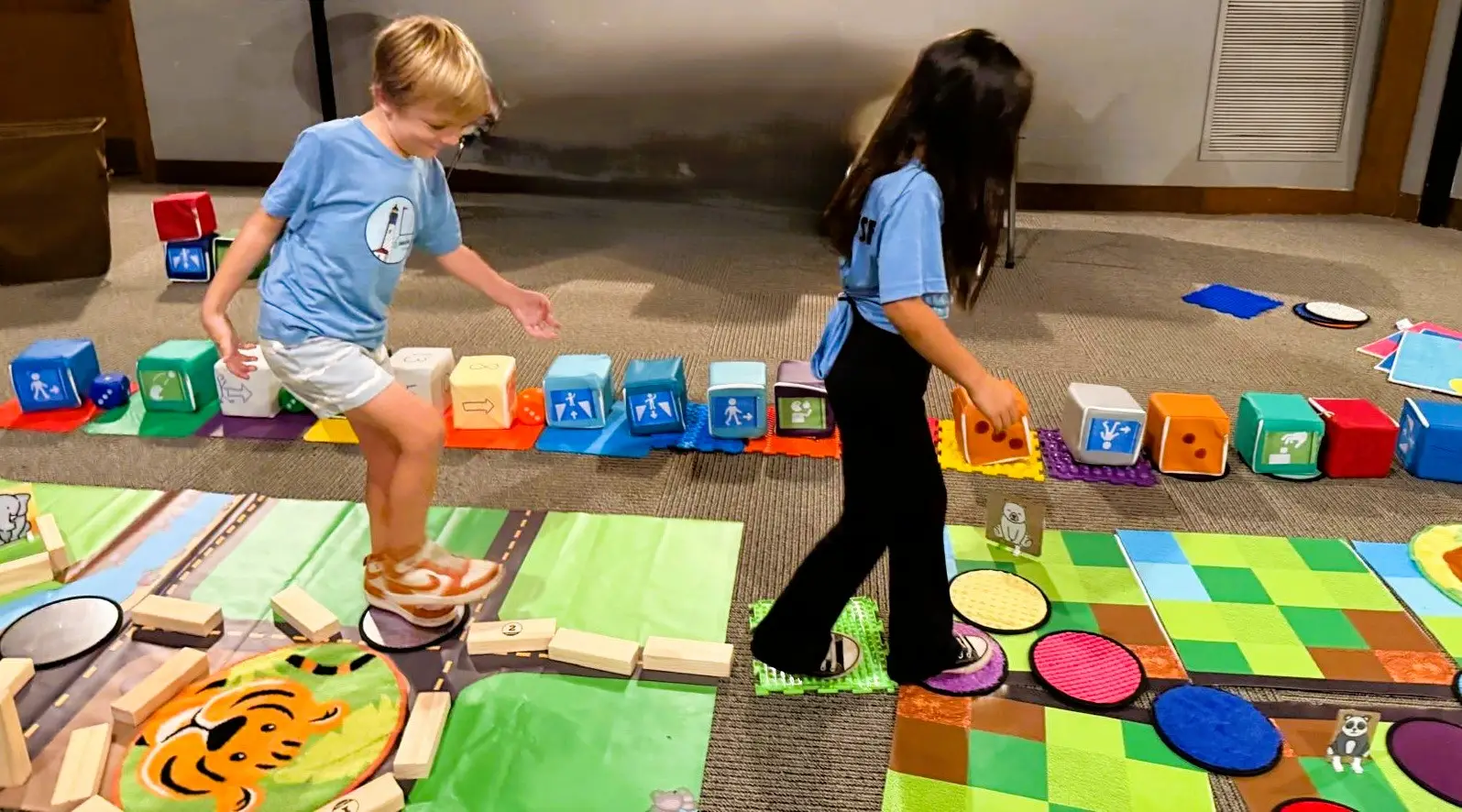 Two children walk along a story-coded floor path, following a sequence they created using large foam coding blocks. They test the code step-by-step while classmates (not visible) watch for errors to help debug the sequence.