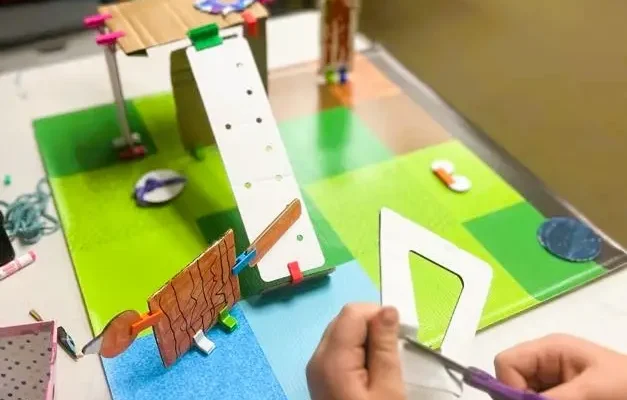 A child cuts a cardboard piece while working on an early-stage animal habitat model. The prototype includes ramps, platforms, and hand-built structures connected with colorful clips. Materials are spread across the table, showing active testing and adjustment during the hands-on engineering task.