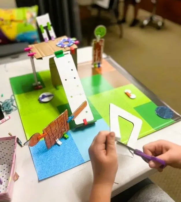A child cuts a cardboard piece while working on an early-stage animal habitat model. The prototype includes ramps, platforms, and hand-built structures connected with colorful clips. Materials are spread across the table, showing active testing and adjustment during the hands-on engineering task.