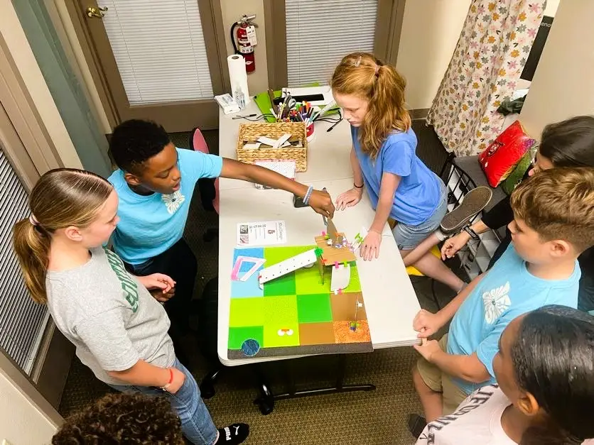 A group of eight upper-elementary students stand around a completed habitat model on a table. One student points to a feature on the model while others watch and listen, engaging in group discussion about the design.