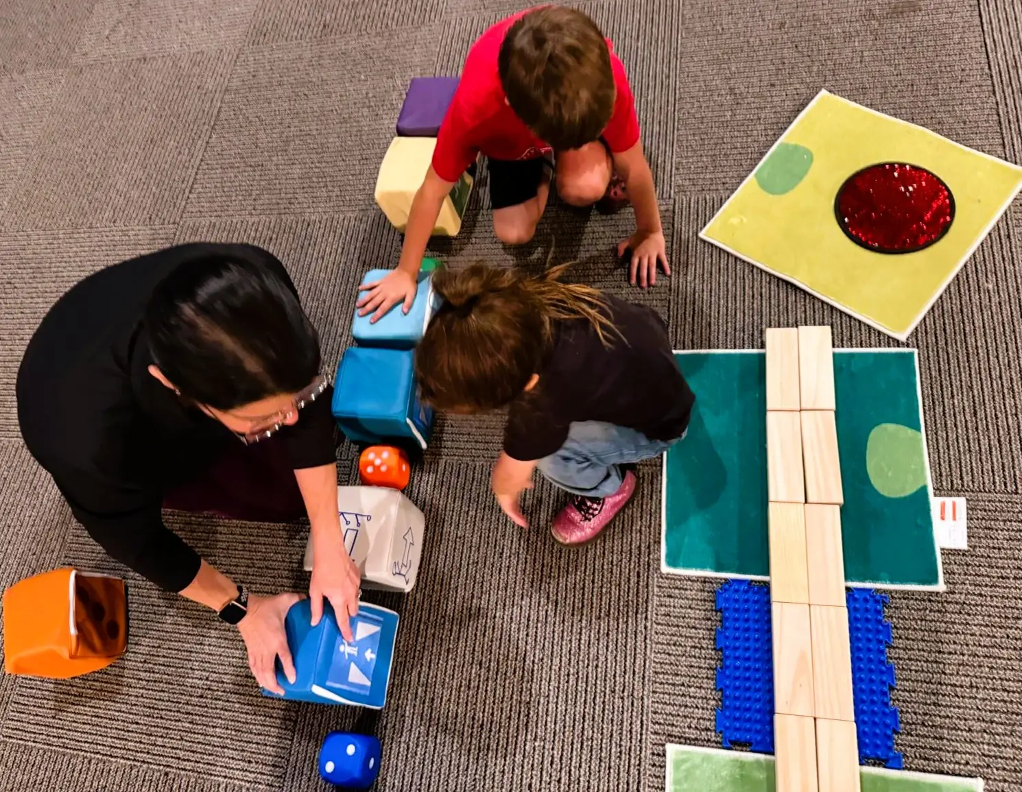 Two young children and an adult arrange large foam cubes with directional arrows on the floor alongside sensory mats and wooden planks, creating a movement-based coding sequence.