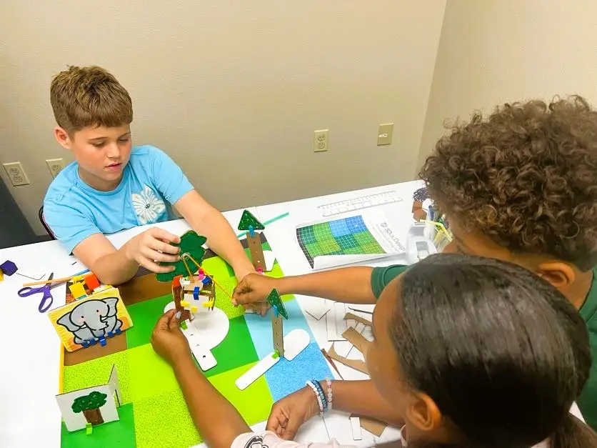 Three upper-elementary students work together on a pulley system attached to a cardboard habitat model. They adjust string tension and components to lift the elephant’s food toward a tree platform.