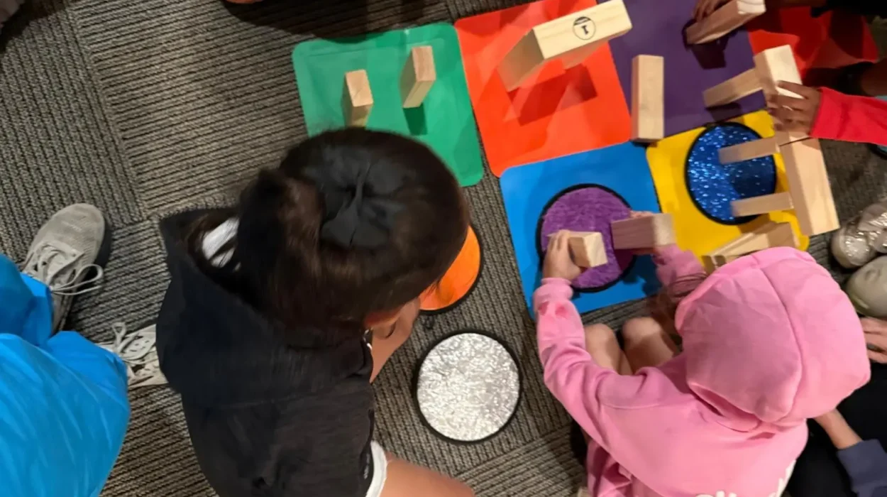 Several children sit on the floor building structures with wooden blocks on colorful tactile mats, adjusting and repositioning pieces while working together.