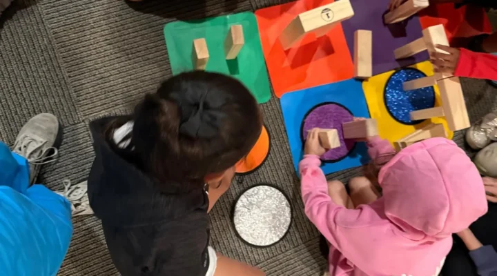 Several children sit on the floor building structures with wooden blocks on colorful tactile mats, adjusting and repositioning pieces while working together.