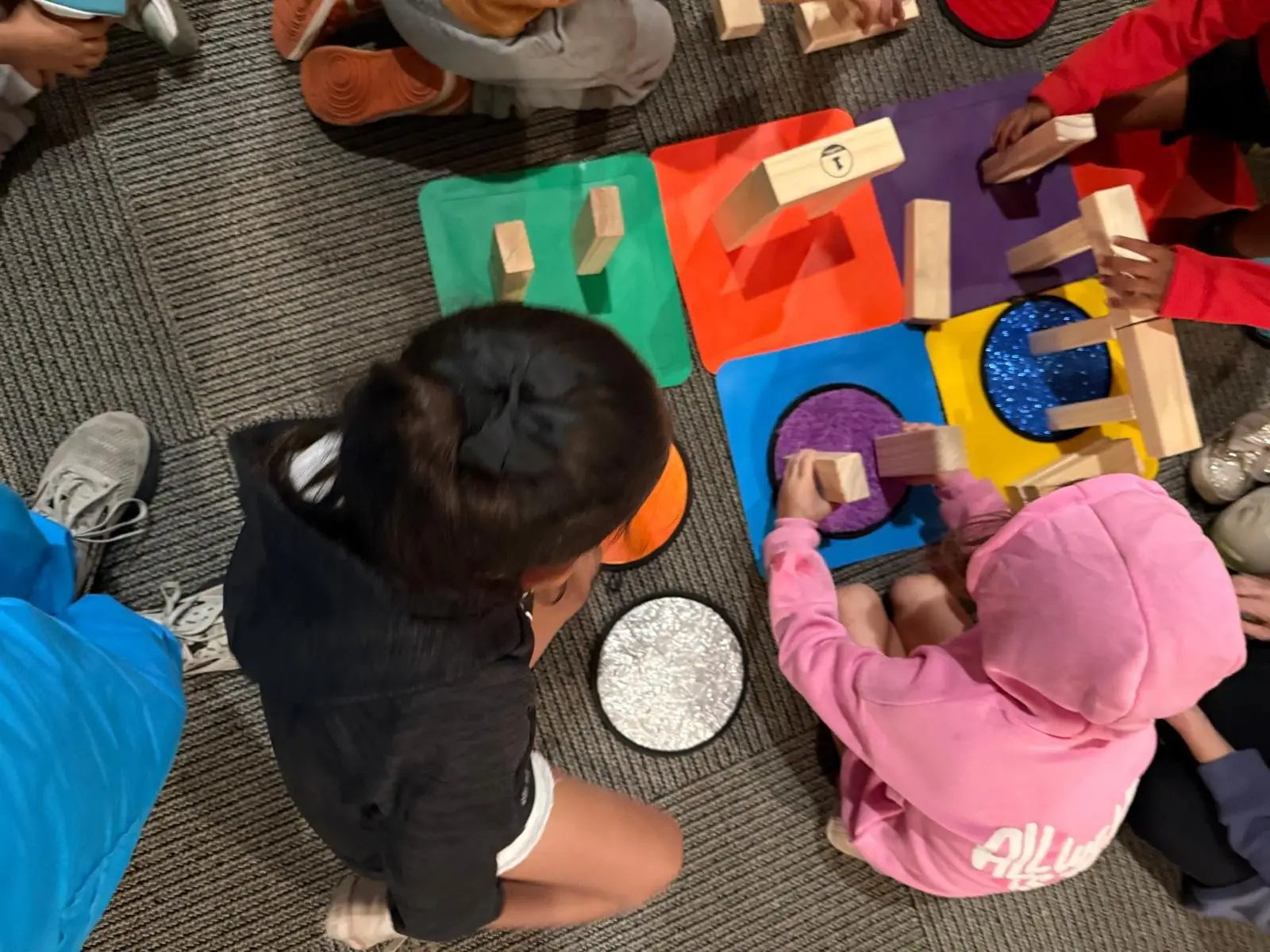 Several children sit on the floor building structures with wooden blocks on colorful tactile mats, adjusting and repositioning pieces while working together.