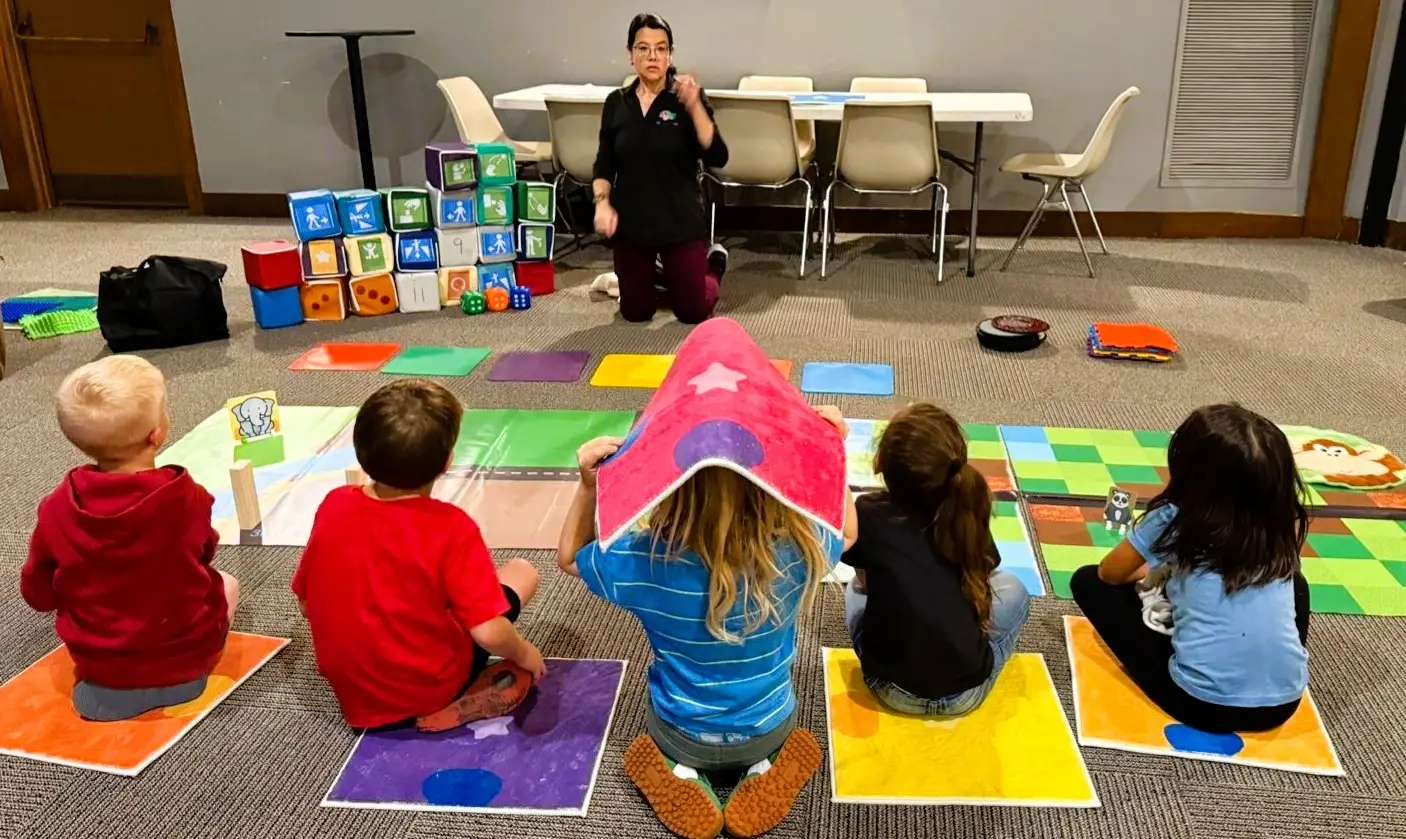 A facilitator kneels in front of a group of young children seated on individual colored mats, introducing the next activity while coding cubes and materials are displayed behind her.